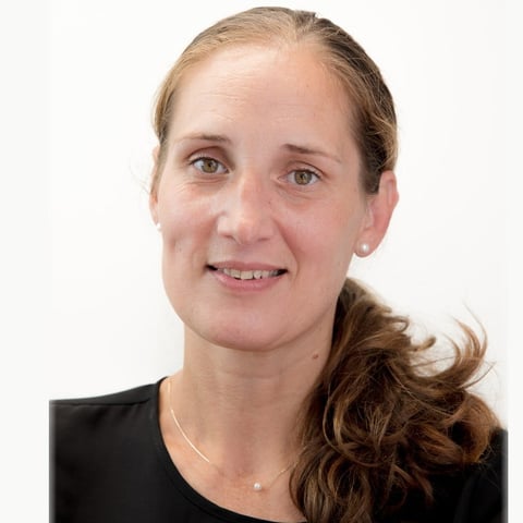 Portrait of a woman with light brown hair in a ponytail, wearing a black top and necklace, smiling at the camera against a white background