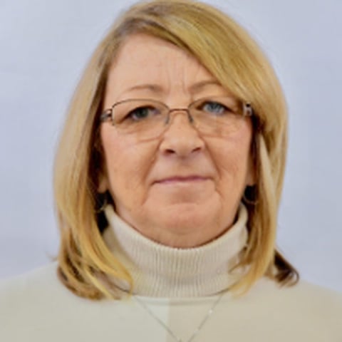 Portrait of a woman with shoulder-length blonde hair wearing glasses and a cream-colored turtleneck sweater against a light blue background