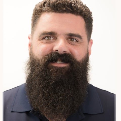 Headshot of a man with a full dark beard and styled hair wearing a dark blue jacket against a white background.
